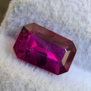 Puede incluir: Una piedra preciosa rectangular facetada en un tono magenta rosado vibrante y translúcido. La piedra presenta bordes afilados y una superficie pulida, con inclusiones oscuras e irregulares visibles dentro de su estructura cristalina.