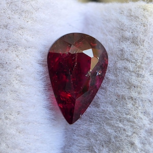 Puede incluir: Una piedra preciosa facetada en forma de pera con un color rojo profundo y translúcido. La piedra presenta múltiples facetas triangulares y trapezoidales que reflejan la luz, creando una estructura interna compleja con inclusiones visibles y tonos rojos.