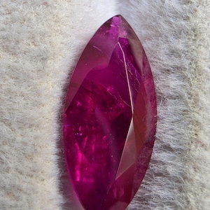 Puede incluir: Una piedra preciosa de corte marquesa con un color magenta rosado profundo y vibrante. La piedra presenta una superficie facetada que crea reflejos de luz geométricos y claridad interna, mostrando una forma ovalada puntiaguda.