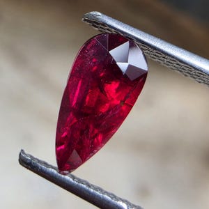 以下が含まれることがあります： 金属製のピンセットで保持された、ファセットカットが施された洋梨型の深い赤色の宝石。石には内部のインクルージョンが見られ、鮮やかな深紅の色合いを呈しており、光がその幾何学的なファセットに反射して輝いています。