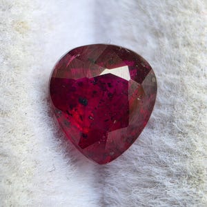 May include: A heart-shaped faceted gemstone in a deep, vibrant magenta-red color. The stone features a polished surface with numerous geometric facets and visible dark, speckled inclusions throughout its translucent, crystalline structure.