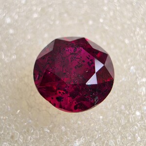 May include: A round, faceted gemstone in a deep, saturated magenta-red color. The stone features a brilliant cut with multiple triangular and kite-shaped facets that reflect light, and it contains visible dark, irregular internal inclusions.
