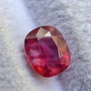 May include: A faceted, cushion-cut gemstone displaying a vibrant gradient of deep pink and red hues. The stone features a polished surface with geometric facets that reflect light, creating internal highlights and shadows across its translucent body.