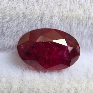 Puede incluir: Una piedra preciosa de forma ovalada en color rojo intenso con una superficie facetada. La piedra muestra un tono carmesí rico y saturado con múltiples cortes geométricos que reflejan la luz sobre su cuerpo pulido y translúcido.
