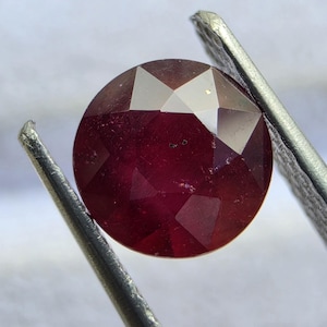 Puede incluir: Una piedra preciosa redonda y facetada de color rojo intenso sostenida por unas pinzas de metal. La piedra presenta una talla rosa con facetas triangulares que reflejan la luz, mostrando un tono carmesí intenso con inclusiones internas.