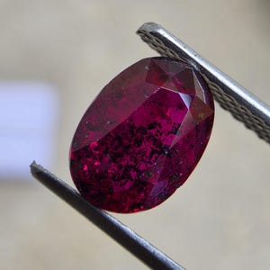 May include: An oval-cut, deep magenta gemstone with prominent dark, irregular inclusions throughout its translucent body. The stone features a faceted surface that reflects light and is held securely by a pair of metal tweezers.
