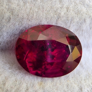 Puede incluir: Una piedra preciosa de color rojo intenso y forma ovalada con una superficie facetada. La piedra muestra un color carmesí rico y saturado con inclusiones internas y cortes geométricos nítidos que reflejan la luz en su superficie.