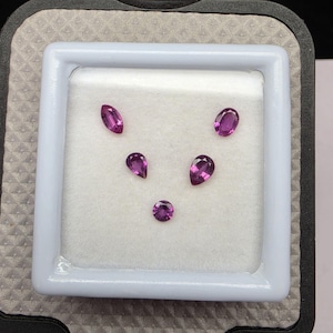 May include: Five faceted loose gemstones in vibrant shades of magenta and purple, arranged in a V-shape. The collection includes one marquise cut, one oval cut, two pear-shaped stones, and one round cut gemstone, all displayed on a white foam insert.