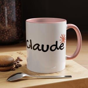 May include: A white ceramic mug with a soft pink interior and handle. The exterior features the name Claude written in black script lettering, accompanied by a small orange starburst graphic positioned above the letter e.