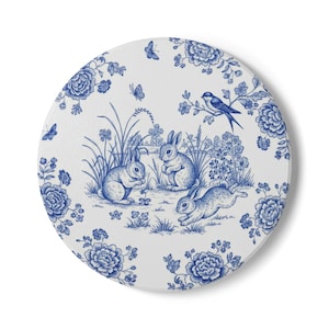 May include: Round white decorative plate with a blue floral and bunny design. The plate features a detailed illustration of rabbits, flowers, and a bird. The border is adorned with blue floral patterns and butterflies.