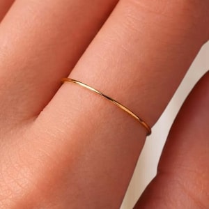 May include: A thin, minimalist gold band ring worn on a finger. The ring features a smooth, polished, and circular design with a simple, continuous metallic finish that encircles the finger, showcasing a clean and understated aesthetic.