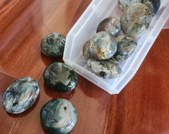 Op de afbeelding: Een verzameling groene, grijze en witte agaatstenen in een doorzichtige plastic container. De stenen zijn glad en gepolijst en hebben verschillende patronen en texturen.