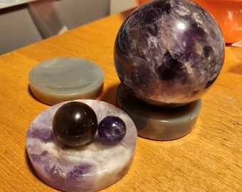 May include: Three amethyst crystal spheres on stands. The largest sphere is purple with white swirls and is on a gray stand. The two smaller spheres are on a purple stand with white swirls. One sphere is black and the other is purple.