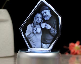 Puede incluir: Una pantalla de fotos de cristal personalizada con una imagen en blanco y negro de una pareja con tazas. El cristal en forma de diamante se asienta sobre una base plateada con una luz brillante. El fondo es negro.