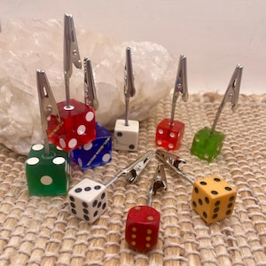 May include: A collection of memo holders featuring colorful dice bases in red, green, blue, white, and yellow. Each die is attached to a silver-toned metal alligator clip on a thin wire stem, designed to hold small cards or notes upright.
