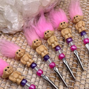 May include: A collection of six vintage-style hair clips. Each clip features a small, tan troll doll with pink hair, adorned with purple and pink beads. The clips have a silver-colored metal finish and are arranged on a textured surface.