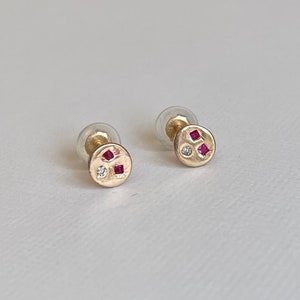 Puede incluir: Un par de pendientes de oro con un diseño redondo texturizado. Cada pendiente tiene tres pequeñas piedras preciosas: dos rojas y una blanca.