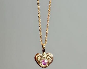 Könnte beinhalten: Eine goldfarbene Halskette mit herzförmigem Anhänger. Der Anhänger hat eine strukturierte Oberfläche und einen kleinen, runden, rosa Edelstein in der Mitte. Die Kette ist filigran und gedreht.
