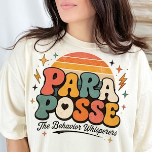 May include: Cream-colored t-shirt featuring the text "PARA POSSE" in colorful, retro-style lettering with a heart and a sunset design. Below the text, it reads "The Behavior Whisperers". The shirt has short sleeves.