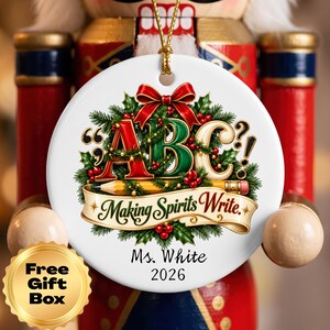 May include: A round white ceramic ornament featuring a green wreath with a red bow, a yellow pencil, and the text ABC and Making Spirits Write. Below the wreath, the text reads Ms. White and 2026. A gold string is attached to the top.