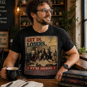 May include: Black t-shirt with a vintage-style graphic featuring the text "GET IN, LOSERS.. WE'RE SIGNING THE DECLARATION" and an illustration of historical figures. The person is holding a black coffee mug and sitting at a wooden table.