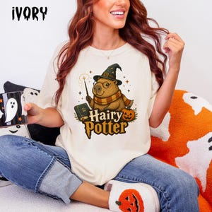 May include: An ivory t-shirt featuring a graphic of a capybara wearing a wizard hat and scarf, holding a wand next to a book and pumpkin. The text "Hairy Potter" is printed below in a stylized font. The shirt is paired with blue jeans.