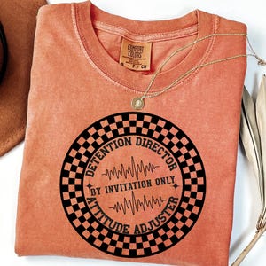 May include: A burnt orange t-shirt with a black and white checkered circle graphic that says "Detention Director By Invitation Only Attitude Adjuster".