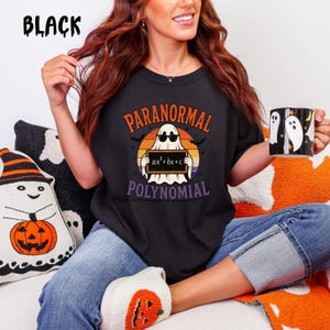 May include: A black t-shirt featuring a graphic of a ghost wearing sunglasses and holding a chalkboard with the equation ax²+bx+c written on it. The text PARANORMAL and POLYNOMIAL surrounds the ghost, set against an orange and purple sunset.