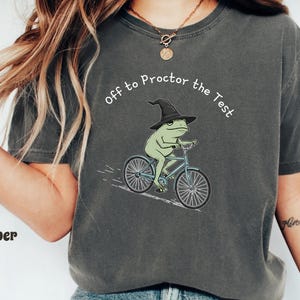 May include: Dark gray t-shirt featuring a green frog wearing a witch hat and riding a blue bicycle. The text "off to Proctor the Test" is arched above the frog. The shirt is a casual, unisex style.