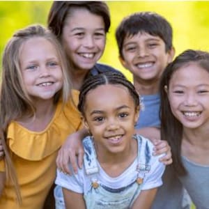 Può includere: Un gruppo di cinque bambini sorridenti di diverse etnie e generi, che posano vicini all'aperto. I bambini indossano top casual di vari colori, tra cui giallo, blu e bianco. Lo sfondo è verde sfocato.