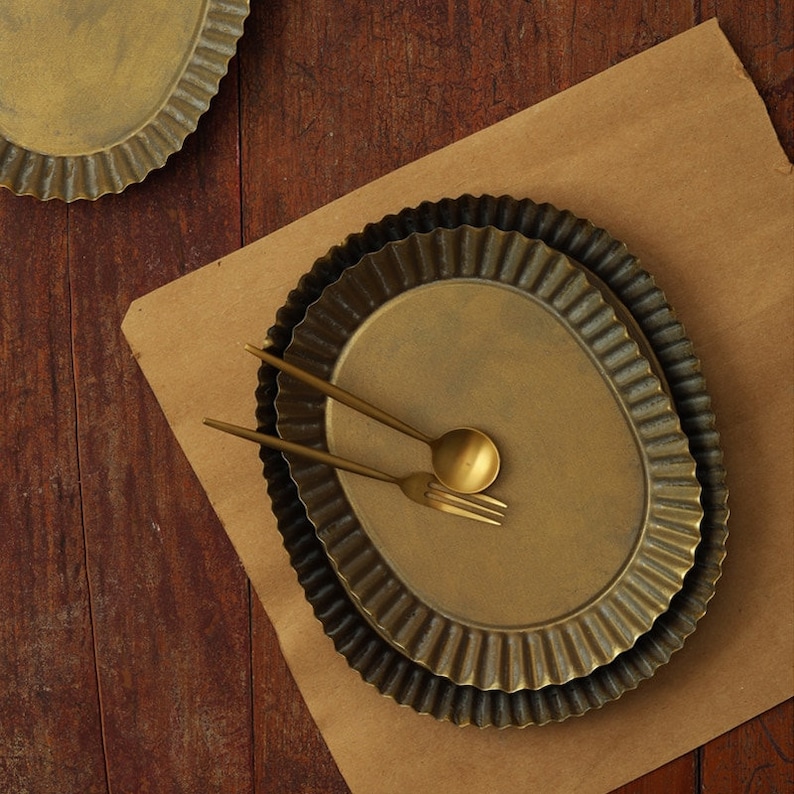 May include: Two gold metal pie plates with fluted edges, stacked on top of each other. A gold fork and spoon are placed on top of the plates.