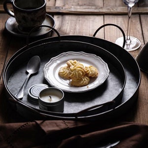 May include: Three black metal serving trays with a white plate with cookies on the top tray. A silver spoon and a small candle are on the tray.