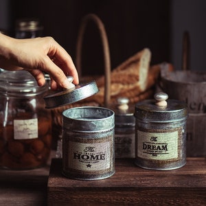 May include: Three metal canisters with lids, each featuring a different design. The canisters are labeled "The Home", "The Dream", and "The Travel".