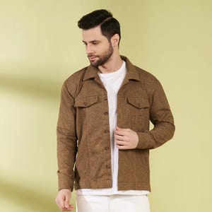 May include: A brown button-up jacket with two chest pockets, worn over a white t-shirt. The outfit includes white jeans and black and white sneakers. The background is a solid light green.