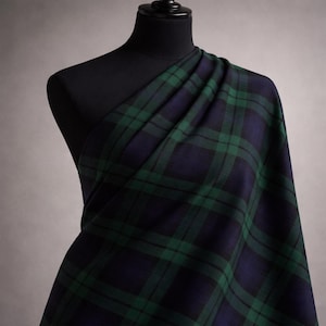 May include: A length of fabric featuring a green and navy blue tartan pattern, draped over a black mannequin. The fabric's design consists of intersecting lines forming a grid. The material's texture and drape are highlighted, showcasing the traditional Scottish design.