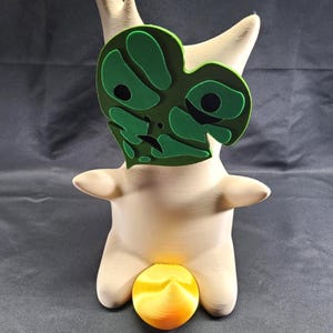 May include: A beige, stylized figurine with a green face and an orange sphere. The figurine has a tree-like appearance with a layered green face, two arms, and a rounded base. The orange sphere is positioned below the figurine.