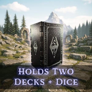May include: A black card deck box with silver accents and the text "HOLDS TWO DECKS + DICE". The box features a textured surface and a silver emblem. The background shows a scenic outdoor landscape with mountains and trees.