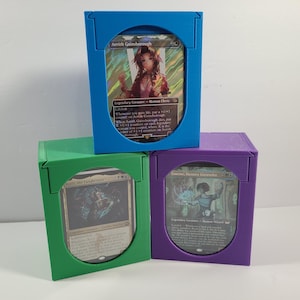May include: Three colored card storage boxes in blue, green, and purple, each holding a Magic: The Gathering card. The blue box displays a card with a woman in a red outfit. The green box shows a card with a dark figure. The purple box displays a card with a person in blue.
