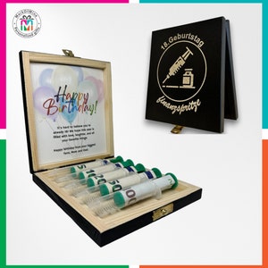 May include: A wooden gift box with a hinged lid, containing syringes filled with rolled-up currency. The box has a printed birthday message and a separate black sign with German text and a syringe graphic. The syringes have green plungers.
