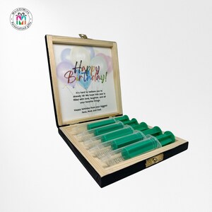 May include: A wooden box with a hinged lid, containing six green and clear syringes. The box has a light wood frame and a black interior. A framed print inside the lid reads "Happy Birthday!" with a balloon graphic.