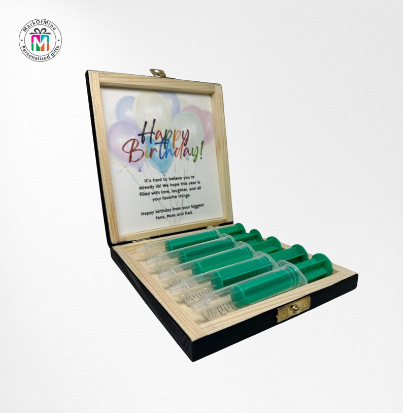 May include: A wooden box with a hinged lid, containing six green and clear syringes. The box has a light wood frame and a black interior. A framed print inside the lid reads "Happy Birthday!" with a balloon graphic.