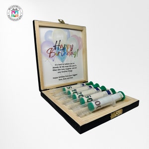May include: A wooden box with a black frame and a light wood interior. Inside, a card displays balloons and "Happy Birthday!". Several syringes are filled with rolled-up banknotes. A personalized gift, ideal for a special birthday.