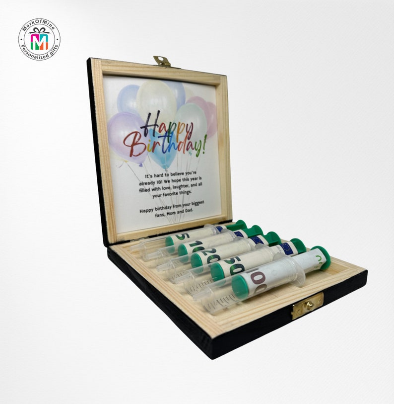 May include: A wooden box with a black frame and a light wood interior. Inside, a card displays balloons and "Happy Birthday!". Several syringes are filled with rolled-up banknotes. A personalized gift, ideal for a special birthday.