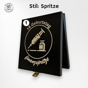 May include: A black wooden sign with a hinged stand. The sign features a circular design with a syringe and vial, and the text "18. Geburtstag finanzspritze". The sign also has the text "Stil: Spritze".