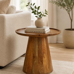 Puede incluir: Una mesa auxiliar redonda de madera con una base de pedestal cónica y una parte superior con borde elevado. Sobre la mesa hay dos libros apilados titulados The Kinfolk Home y Live Beautiful, coronados por un pequeño jarrón de cerámica beige con hojas verdes.