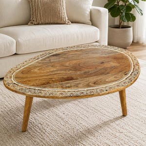 Op de afbeelding: Een ovale salontafel met een oppervlak van natuurlijk houtnerf en een decoratieve rand met witte en bruine geometrische patronen. De tafel wordt ondersteund door drie taps toelopende houten poten en heeft een gladde afwerking.