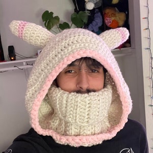 May include: A white crocheted hooded hat with pink trim and a matching white crocheted neck warmer. The hat has two bunny ears.