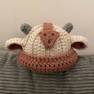 Könnte beinhalten: Ein handgefertigter Häkelhut in Form einer Zeichentrickfigur. Der Hut hat eine cremefarbene Basis mit einem rostfarbenen Rand und Akzenten. Er hat graue Hörner und rostfarbene Details, die an den Kopf einer Figur erinnern.