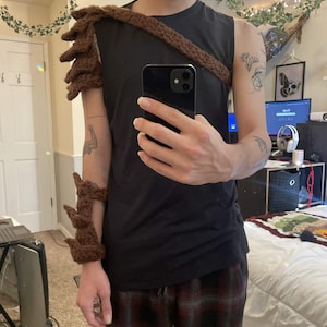 Könnte beinhalten: Ein braunes gehäkeltes Schulter- und Armteil mit einem stacheligen Design. Das Armteil umschließt den Unterarm und das Handgelenk. Das Schulterteil ist durch einen Riemen, der über die Brust verläuft, mit dem Armteil verbunden.