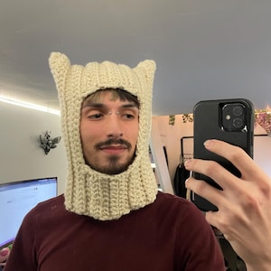 May include: A cream-colored, crocheted balaclava with pointed ear details. The head covering is made of thick yarn and features a ribbed neck and face opening. The balaclava is worn by a person in a maroon shirt.